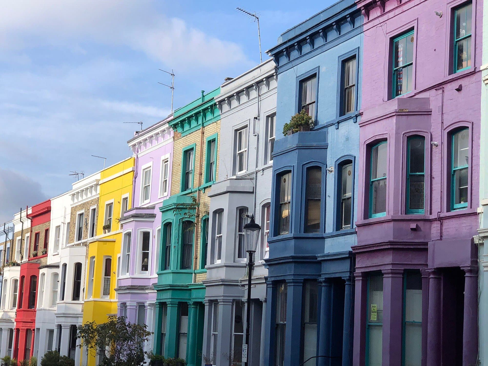 Colorful Victorian-style row houses in Notting Hill, West London. The image showcases a vibrant street with houses painted in bright hues of red, yellow, purple, green, blue, and lavender, creating a picturesque and lively neighborhood scene under a partly cloudy sky. Perfect for promoting travel, architecture, and the unique charm of London's Notting Hill district.