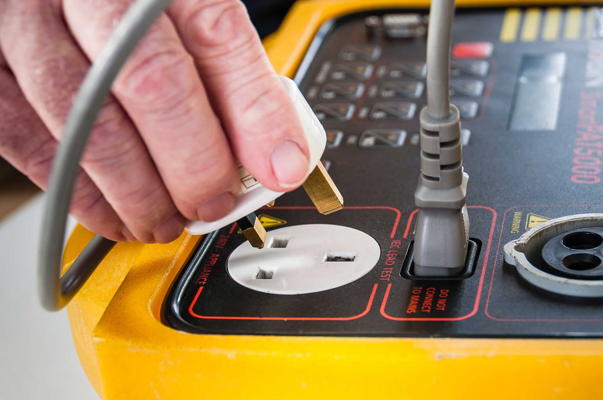 Close-up of an electrical safety test being conducted using a yellow PAT tester device, with a technician inserting a 3-pin plug for appliance compliance verification, emphasising professional PAT testing services.