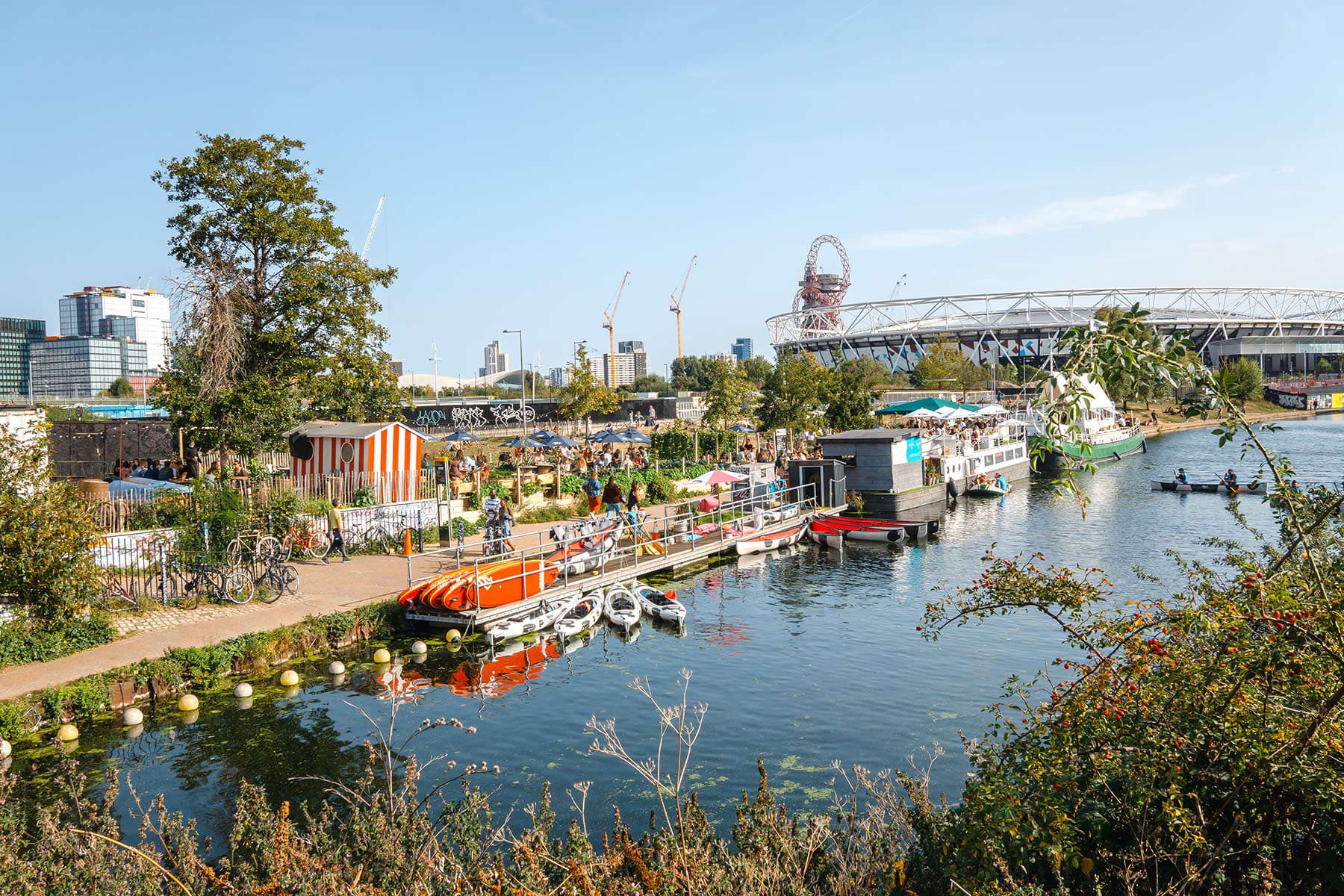 Vibrant scene at Queen Elizabeth Olympic Park in Stratford, East London, featuring the London Stadium, the ArcelorMittal Orbit sculpture, and lively canal activities with boats and visitors enjoying the sunny day.